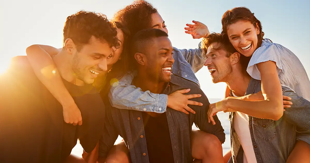 Grupo de jovens adultos, na faixa dos 30 anos, sorrindo e se divertindo juntos ao ar livre sob a luz do sol, representando a importância da comunicação e interação social.