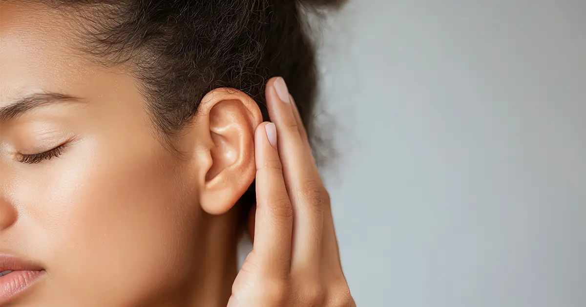 Mulher de perfil com os olhos fechados e a mão atrás da orelha, demonstrando esforço para escutar devido à perda auditiva.