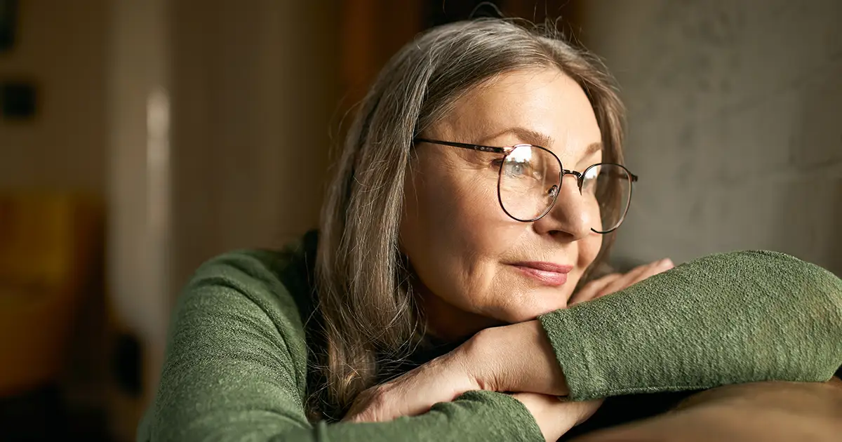 Mulher idosa com óculos de grau, encostada de forma relaxada em um sofá e olhando pensativa pela janela, transmitindo uma sensação de reflexão.